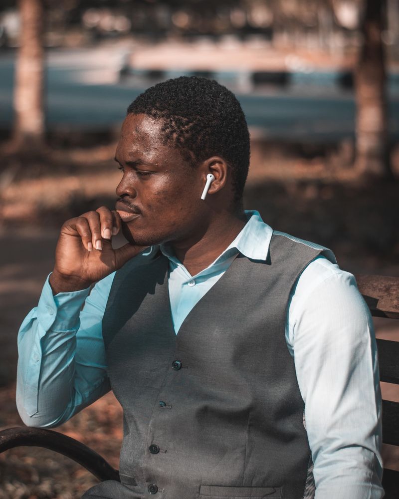 Gift Balogun seated on a park bench, wearing a grey waistcoat over a light blue shirt.